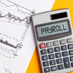 The image has yellow desk with lest side showcasing two papers with graphs with a calculator slightly on top of documents. The calculator shows payroll. Overall, the image depicts the cost of outsourcing payroll services in singapore.